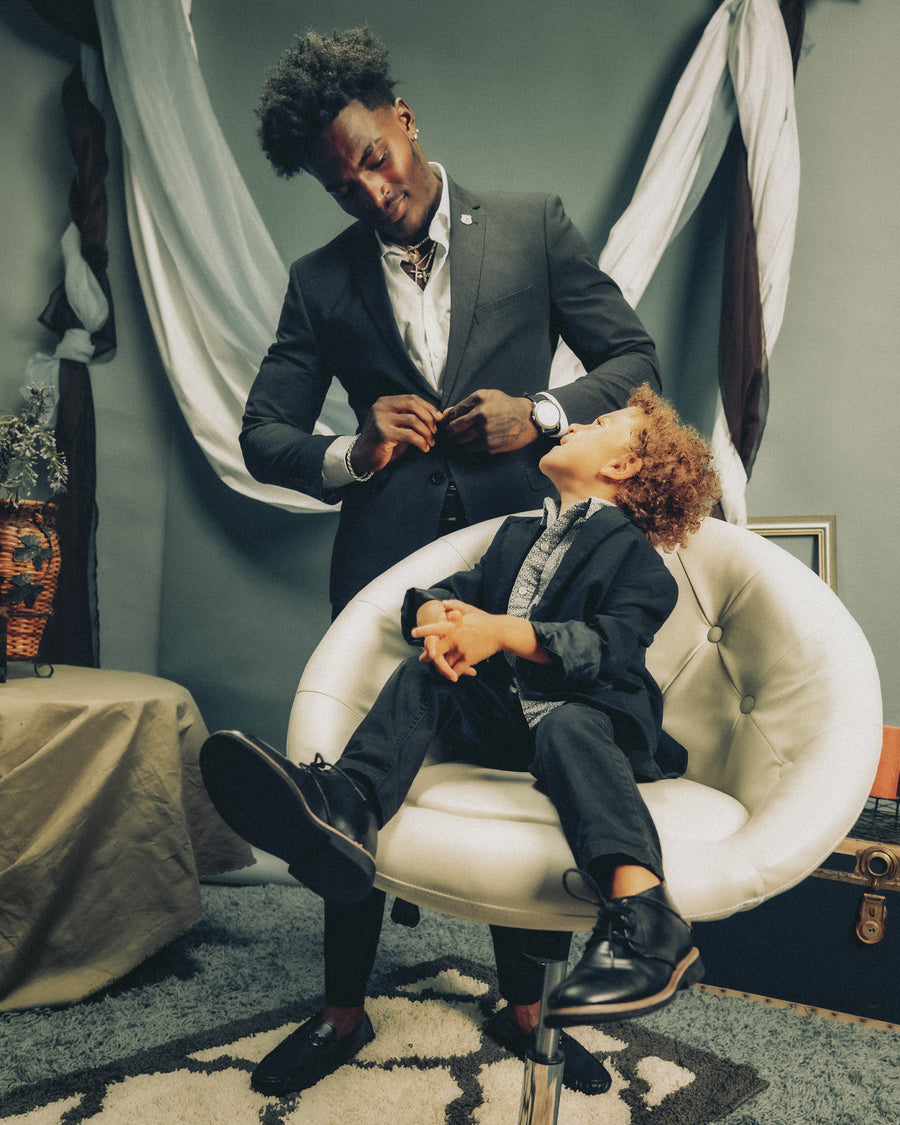 Father and son in matching suits, the boy seated in a white chair