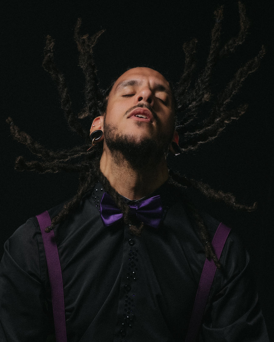 Motion portrait of a man with locs mid-swing, purple bow tie and suspenders